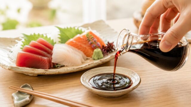 ガラスの醤油さしから小皿に無添加醤油を注ぐ様子。奥には刺身の盛り合わせが置かれた、ナチュラルな食卓風景。